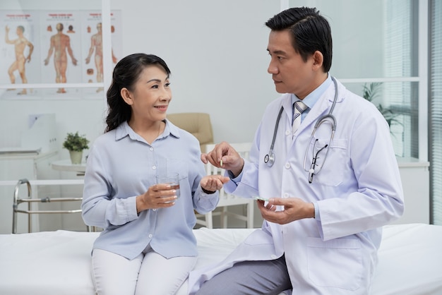 Serious physician giving pills to smiling senior woman sitting on bed with glass of water