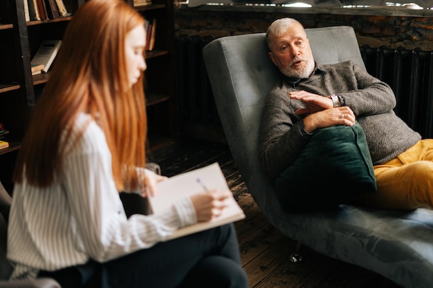 Photo serious red-haired young woman psychologist taking notes in notebook, supporting and consoling depressed older man patient during psychotherapy session.