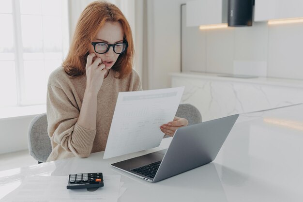 Serious redhead woman focused in paper documents uses calculator and laptop computer