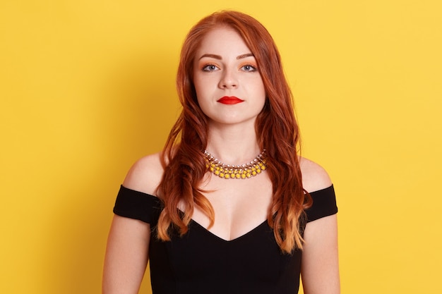 Serious sad ginger female, woman with red lips wearing black elegant dress and golden necklace, posing isolated over yellow wall.