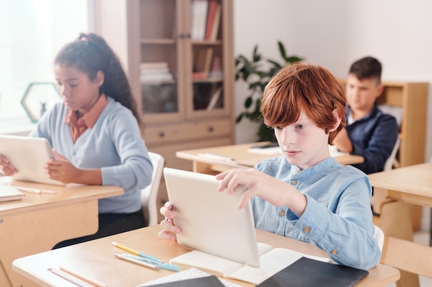 Serious schoolboy with touchpad looking through online document while sitting by desk against intercultural classmates during individual work