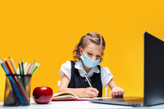 Serious schoolgirl in mask doing her homework with laptop writing in notebook distance education