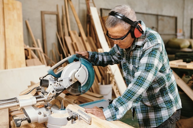 Serious senior Asian man in earmuffs cutting wood plank with circular saw in workshop