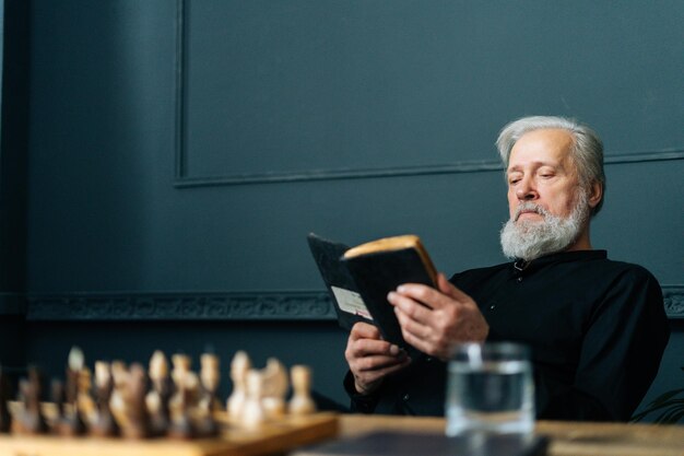 Serious senior grayhaired older male reading paper book at home sitting at wooden table with chess board relax and rest with literature