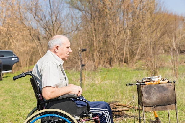 Serious Senior Man Sitting on a Wheelchair at the Park Waiting the Grilled Meat to be Cooked Under the Heat of the Sun.