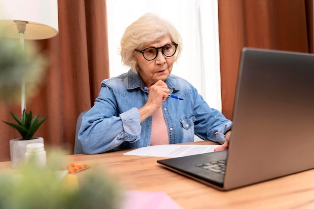 Serious senior woman holding online training while working at home with laptop Old woman wearing eyeglasses teaching students online Beautiful stylish elderly lady browsing site on computer