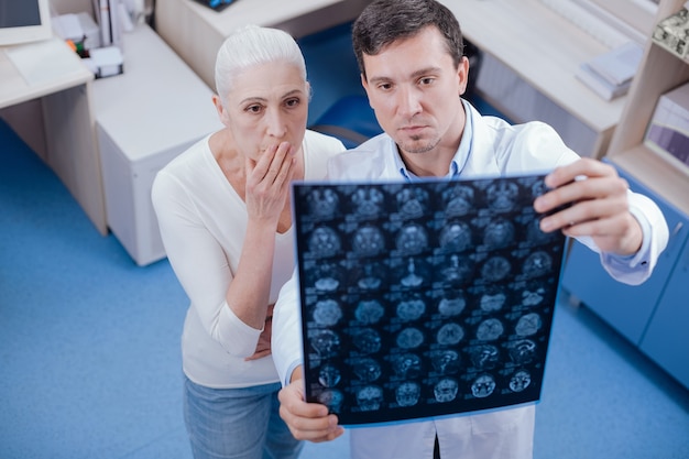 Серьезно потрясенная несчастная женщина смотрит на рентгеновские снимки своего мозга и прикрывает рот, пораженная своим диагнозом
