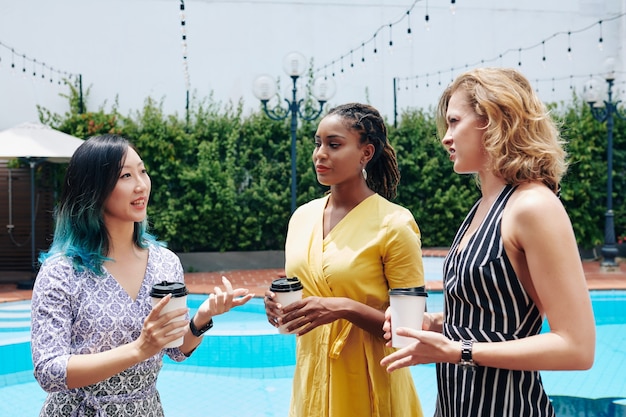 Serious skeptical young businesswomen listening to questionable idea of colleague when standing by swimming pool and having coffee