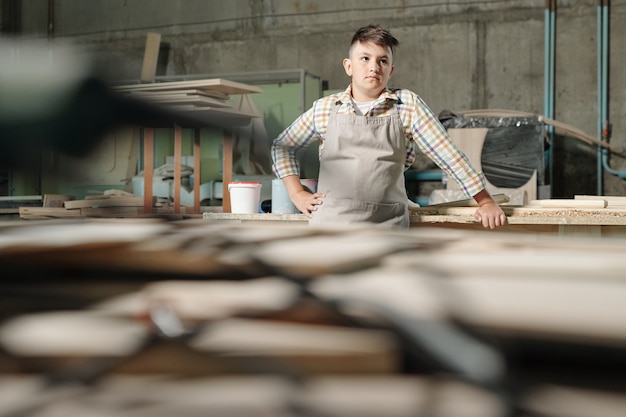 Serious skilled teenage boy in apron and checkered shirt standing at desk in carpentry workshop