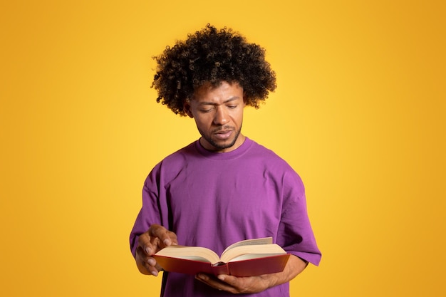 주황색 배경에 고립된 책을 읽는 보라색 티셔츠를 입은 진지한 똑똑한 흑인 성인 곱슬머리 남자