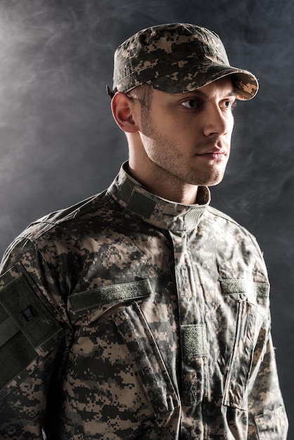 Serious soldier in military uniform and cap on black with smoke