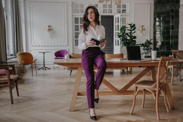 Серьезная испанская деловая женщина в белой рубашке и фиолетовых штанах сидит на столе, смотрит в сторону и держит дневник Целеустремленная брюнетка с распущенными волнистыми волосами планирует свою неделю Деловые люди