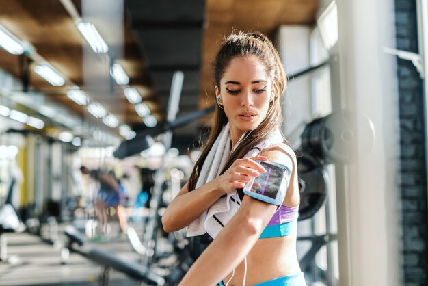 Serious sporty woman in sportswear and towel around neck putting smart phone in phone case on hear arm. In background exercise equipment. Gym interior.