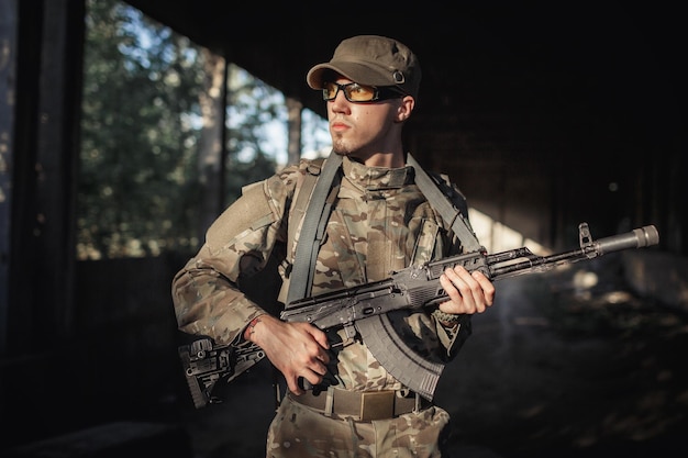 Фото Серьезный суровый солдат в серой камуфляжной форме, в тактических очках и кепке с черным пулеметом в руках внутри большого санкционированного здания