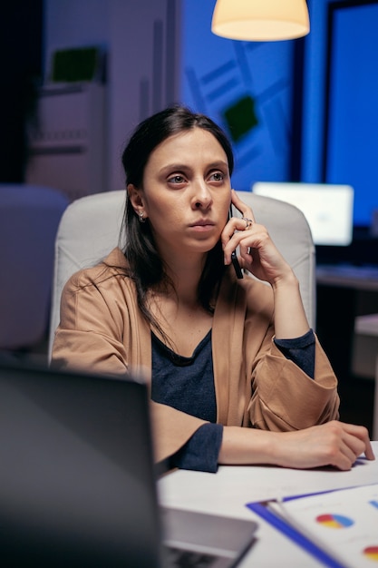 Serious stressed businesswoman having a conversation with a client. Woman entrepreneur working late at night in corporate business doing overtime in the course of phone call.