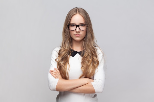 Serious strict woman in glasses standing looking at camera with bossy expression keeps hands crossed