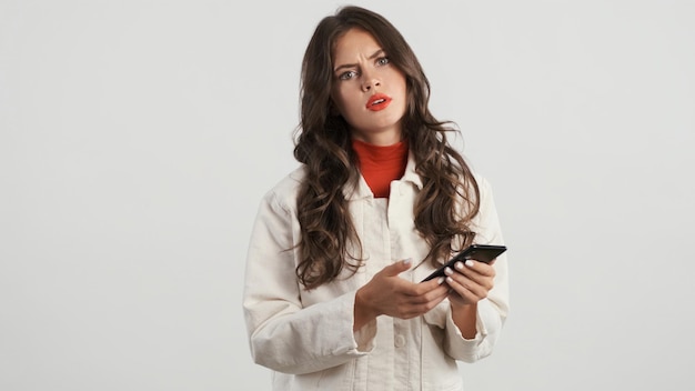 흰색 배경 위에 스마트폰을 사용하여 카메라를 짜증스럽게 보고 있는 진지한 세련된 갈색 머리 소녀