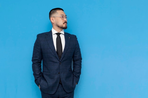 serious successful asian businessman in suit and glasses stands on blue isolated background