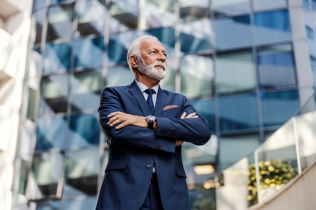 A serious successful bearded senior businessman in a suit is standing in front of his company