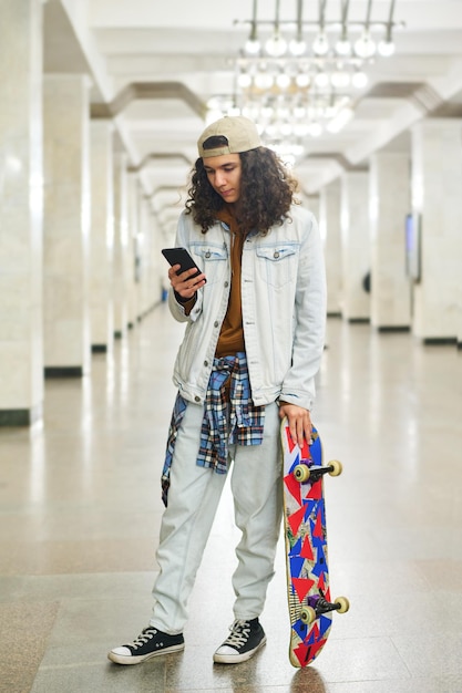 Serious teenage boy with skateboard standing at subway station and texting in smartphone