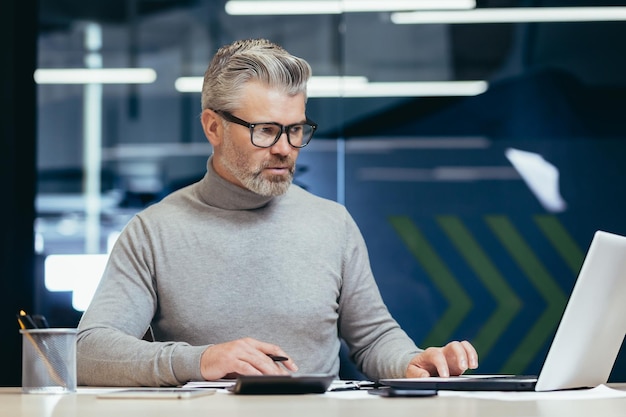 Serious thinking businessman inside office at work with laptop senior gray haired man working