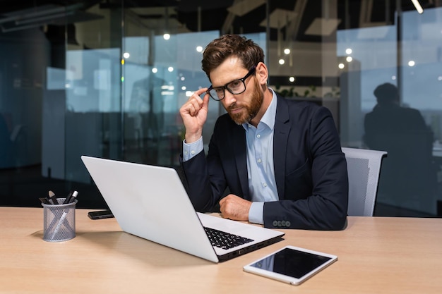 Serious thinking senior businessman working inside office using laptop man in glasses solving