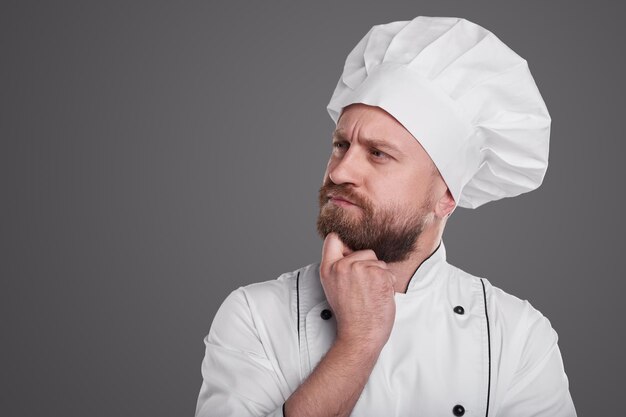 Cuoco maschio barbuto serio e premuroso in uniforme bianca da chef e cappello strofinando il mento e guardando lontano pensieroso su sfondo grigio