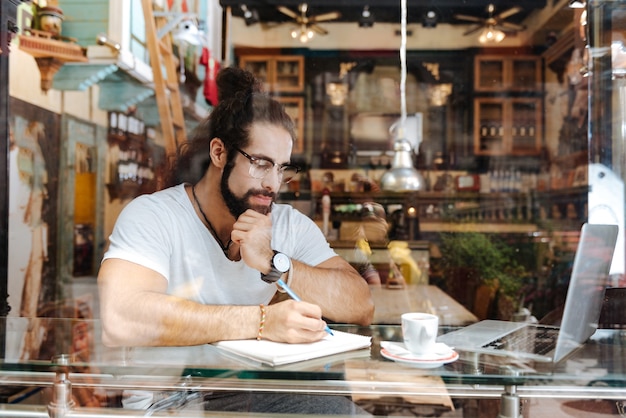 Серьезный вдумчивый человек, сидящий перед ноутбуком во время работы в кафе