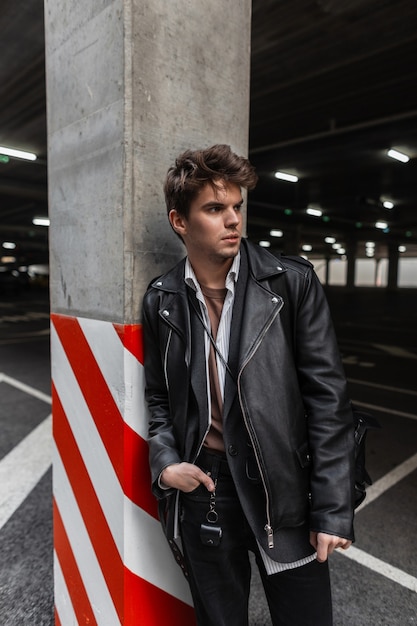 Serious trendy young man model in a youth oversized fashionable black leather jacket with a stylish hairstyle stands near a gray column in a city parking lot. Handsome hipster modern guy in the street