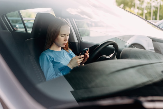 Serious woman holding her smart phone while sitting inside the car.