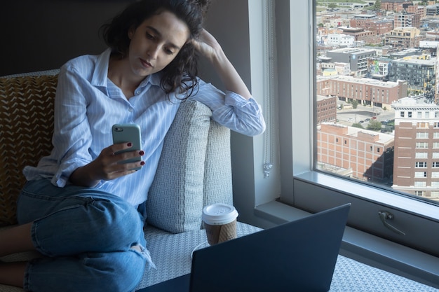 Фото Серьезная женщина, глядя на смартфон во время перерыва в работе, вид на город в фоновом режиме, с копией пространства, концепция домашнего офиса