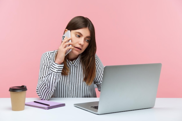 Serious woman office worker talking phone typing on laptop, receiving calls and making online order, coffee cup and notepad lying on table. Indoor studio shot isolated on pink background