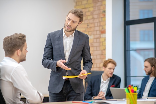 Serious young adult man in business suit with folder standing asks sitting colleague