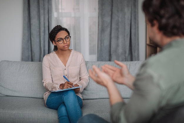 Serious young african american female psychologist in glasses listens to european patient in clinic