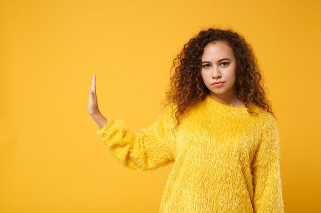 Serious young african american girl in fur sweater posing isolated on yellow orange wall background studio portrait. people lifestyle concept. mock up copy space. showing stop gesture aside with palm