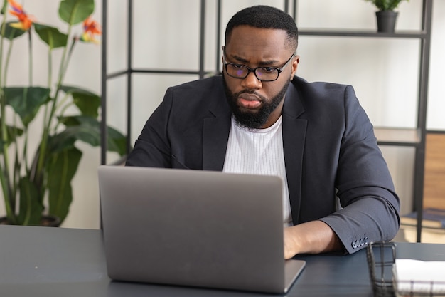 眼鏡をかけた真面目な若いアフリカ系アメリカ人男性は、ラップトップのデスクワークに座っています。ビジネスマンはオンラインでビジネス会議に参加します