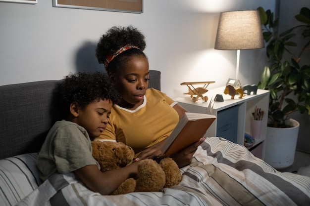 머리 스카프를 두른 심각한 젊은 아프리카계 미국인 어머니는 침대에 누워 피곤한 아들에게 책을 읽어줍니다.