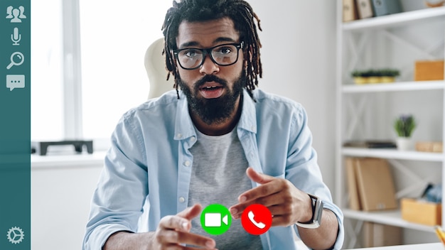 Serious young African man in smart casual wear looking at camera while having video call