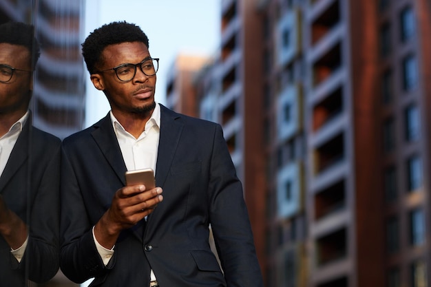 Serious young afroamerican businessman with hipster beard leaning on building wall and texting messa