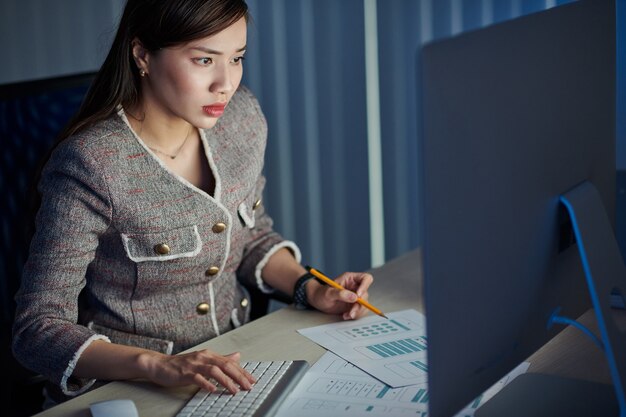 Serious young Asian business lady working in office late at night and making corrections in interface layout for UI designer