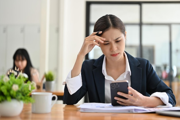 Giovane donna d'affari asiatica seria alla sua scrivania che si occupa di un problema con lo smartphone