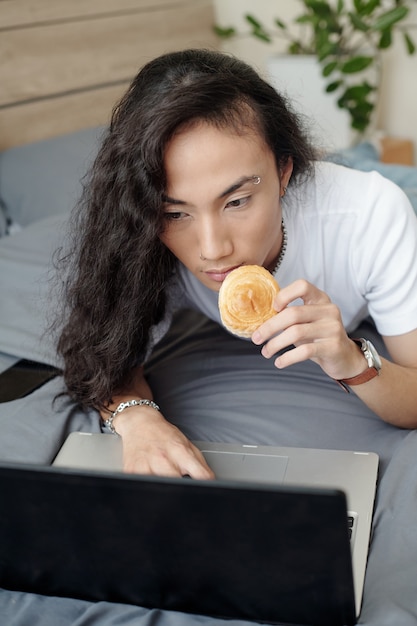 Serious young Asian freelancer with long hair lying on bed and eating bun while using laptop at home