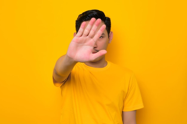 Serious young asian man in casual tshirt makes stop gesture\
covering her face with palm isolated on yellow background people\
lifestyle concept