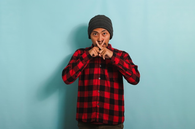 Serious Young Asian man makes a crossed fingers gesture stop talking isolated on a blue background