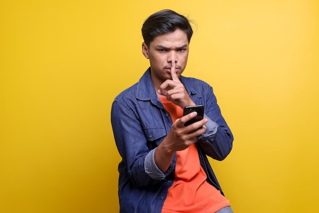 Serious young Asian man showing keep silent gesture while holding smartphone on yellow background