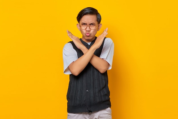 Serious young asian man showing refusal gesture with crossed arms isolated over yellow background