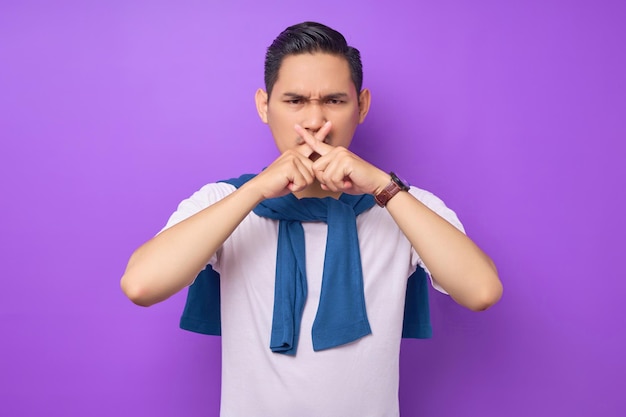 白い t シャツを着て口を覆い、紫色の背景の人々 のライフ スタイル コンセプトに分離された指で交差停止を示す深刻な若いアジア人男性