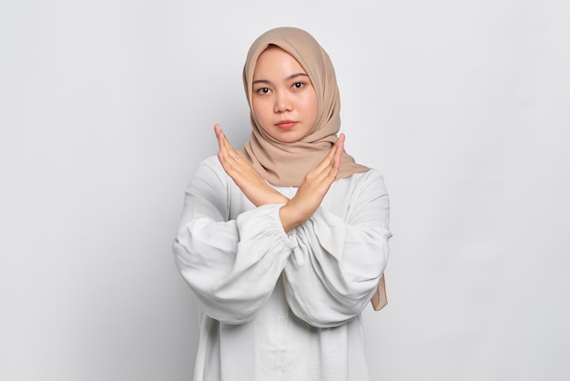 Serious young Asian Muslim woman crossing hands makes stop gesture and demonstrates rejection isolated over white background