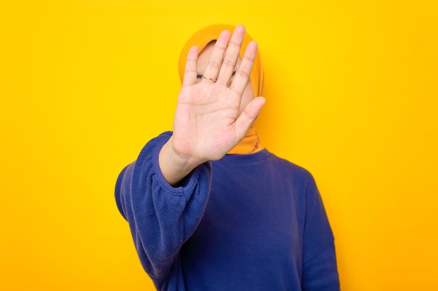 Serious young asian muslim woman dressed in casual sweater\
makes stop gesture and covering her face with open palm isolated\
over yellow background