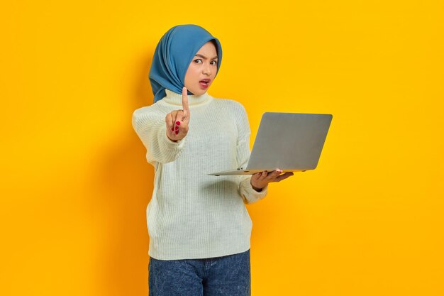 Serious Young Asian muslim woman in white sweater holding laptop and showing stop gesture isolated isolated over yellow background people religious lifestyle concept